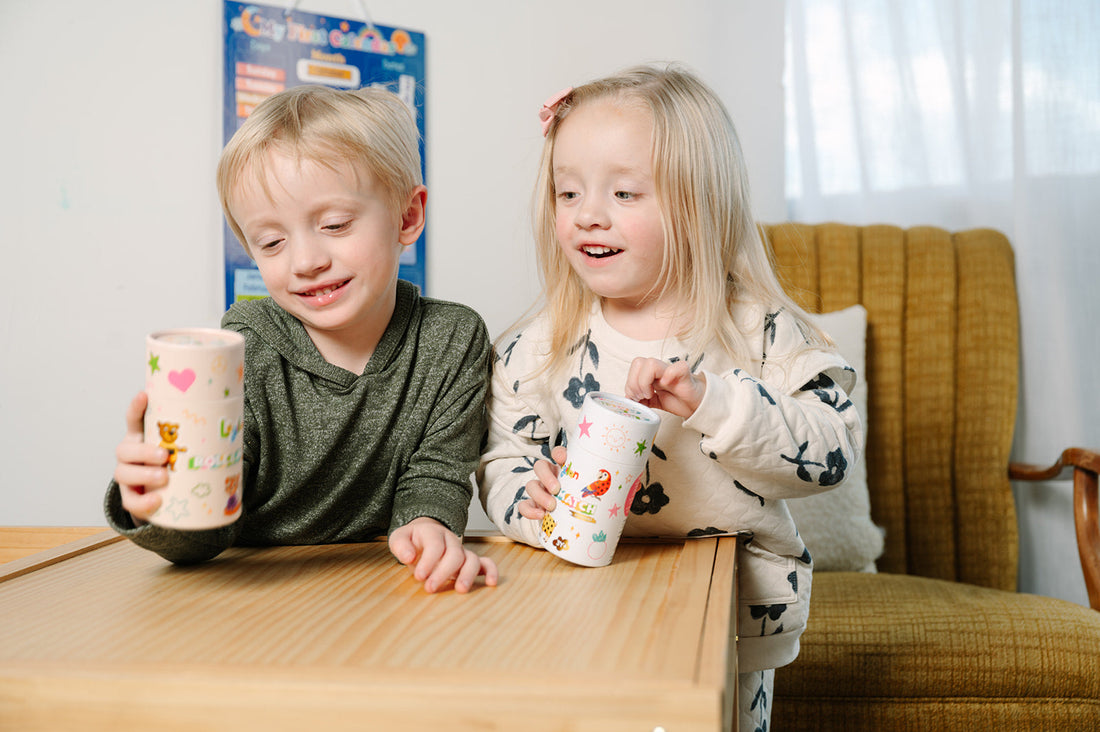 Two children sitting at a table holding colorful cups that contain the game pieces.